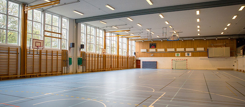 Sporthall med stora fönster, blått golv, ribbstolar, basketkorgar, romerska ringar, mål och läktare.
